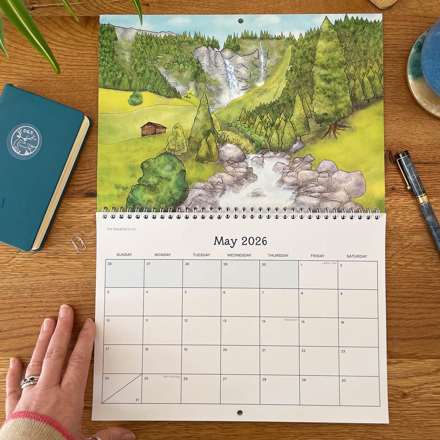 Calendar for May 2026 with a scenic illustration of a landscape on a wooden surface.